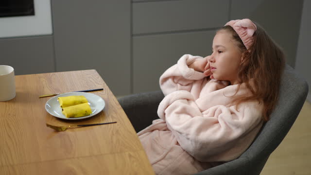 A girl in a bathrobe and a hair band on her head sits on a chair and dreams at the table, on which there is a plate with two filled pancakes.