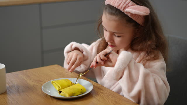 Slow motion. A girl has breakfast with pancakes, sitting indoors in a bathrobe and a hair band on her head. A child eats in the kitchen in a modern apartment.