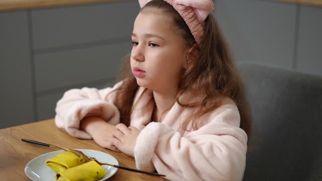 Slow motion. A smiling girl cuts a pancake with filling with a dessert knife, sitting indoors in a bathrobe and a hair band on head. The child lost his appetite when he saw the filling in the pancake.