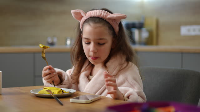 Slow motion. A girl, in a bathrobe and a pink cosmetic hair band with cat ears, sits at the table and eats pancake with filling. The girl looks at the screen of a mobile phone that lies on the table.