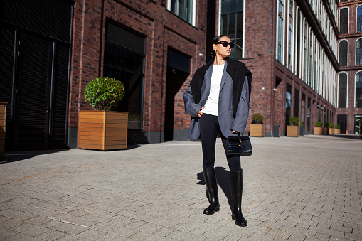 Stylish fashion woman strolls down the street in an oversized blazer, black leggings, and knee high rubber boots. Fashionable outfit, street style.