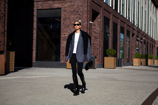 Stylish fashion woman strolls down the street in multilayer clothes oversized blazer, black leggings, and knee high rubber boots. Fashionable outfit, street style.