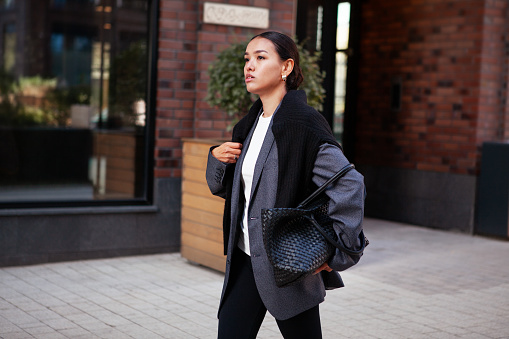 Stylish fashion woman strolls down the street in multilayer clothes oversized blazer, black leggings and large bag. Fashionable outfit, street style.