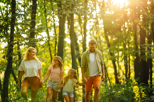 A happy family holding hands and exploring forest while spending sunny summer day in nature. A young parents holding hands with their daughters and spending summer day exploring forest and nature.