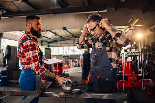 Caucasian men in plaid shirts and work overalls working together in a workshop. Main craftsman is putting his safety headphones and goggles before start grinding while the other one is helping him.