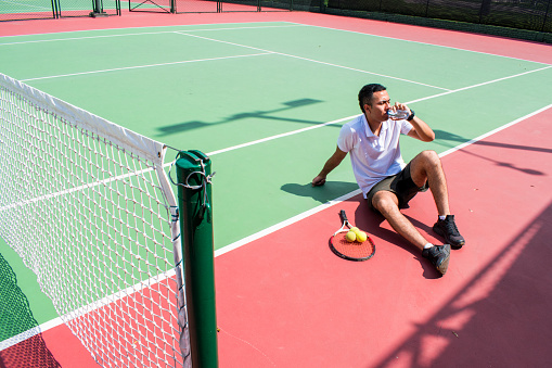 Young latín man tennis player resting and drinking water on tennis court in sunny hot day at summer