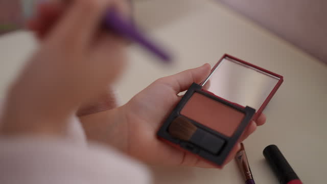 Close-up. Slow motion. An unrecognizable girl holds an open powder compact with a small mirror in her hand and carefully picks up powder with a makeup brush.
