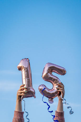 Girl holding 15th birthday balloons over head outdoors, blue sky at background