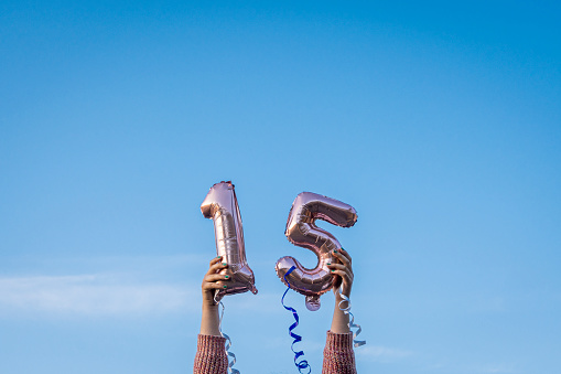 Girl holding 15th birthday balloons over head outdoors, blue sky at background