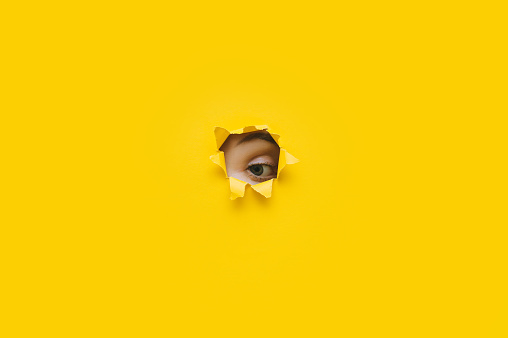 Left eye looking through a hole in a yellow paper. Voyeurism. Woman is watching the husband. A curious look. Jealousy, spying on or overhearing the concept. Copy space.