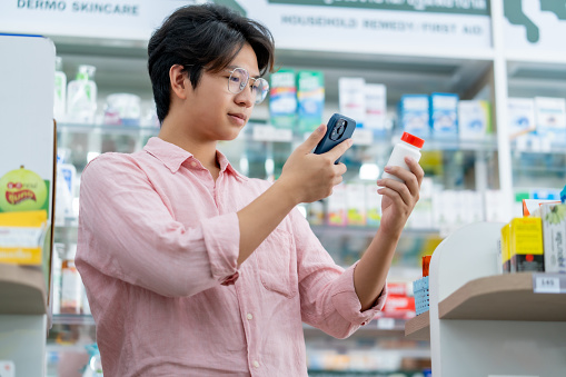 A male customer uses his smartphone to scan a medicine bottle in a pharmacy, comparing products or prices.
