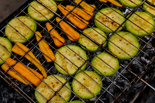 Chopped zucchini and carrots roasting on fire seasoned with aroma herbs and spices. Delicious fresh vegetables grilling on barbecue smoker grid. Diet vegan bbq. Outdoor recreation in backyard