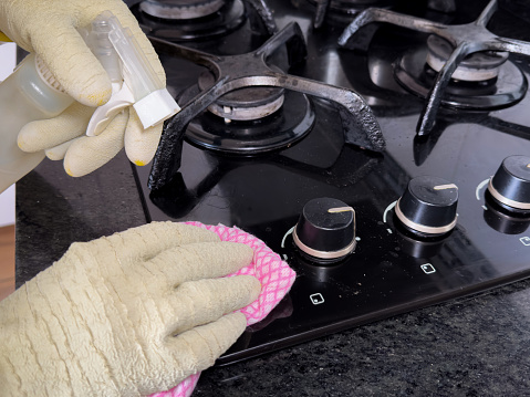A person cleaning a modern black gas stove with a pink cloth and a spray cleaner. The person wears plastic cleaning gloves. Concepts to eliminate fat, cook impeccably and shine your stove
