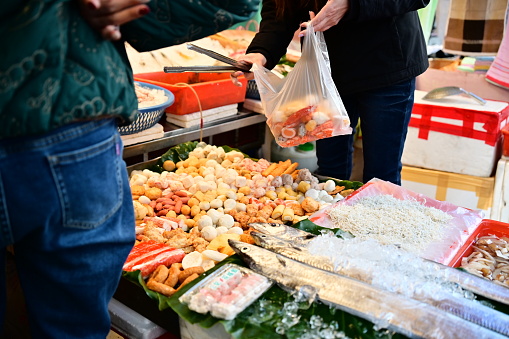 At a market stall, a close-up captures a vendor selling fish and various hot pot ingredients, while a customer is seen selecting items to put in their plastic bag.