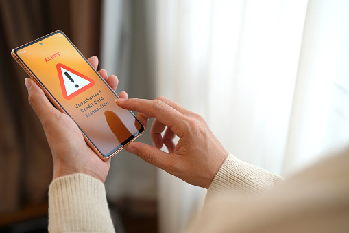 Cropped image of a woman receives an alert message from her bank on her phone, informing her about an unauthorized credit card transaction. Unauthorised use of a transaction refers to the charging of expenses or purchasing of goods and services without the cardholder's consent.