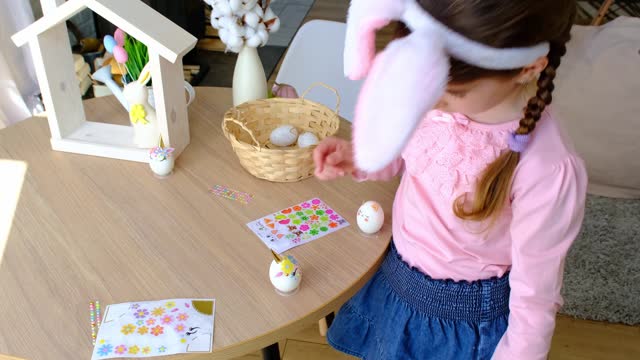 A cute girl with pink bunny ears makes an Easter craft - decorates an egg in the form of a unicorn with rhinestones, horn, flowers in the interior of a house with plants.