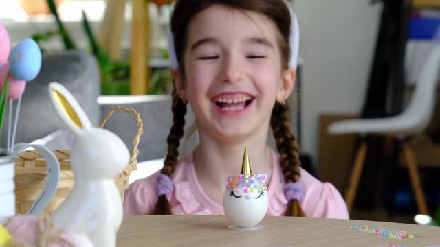 A cute girl with pink bunny ears makes an Easter craft - decorates an egg in the form of a unicorn with rhinestones, horn, flowers in the interior of a house with plants.
