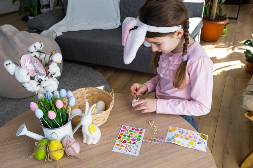 A cute girl with pink bunny ears makes an Easter craft - decorates an egg in the form of a unicorn with rhinestones, horn, flowers in the interior of a house with plants.