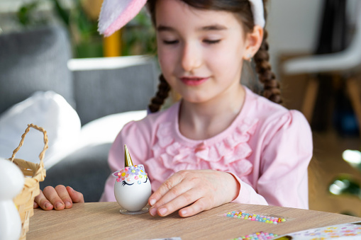 A cute girl with pink bunny ears makes an Easter craft - decorates an egg in the form of a unicorn with rhinestones, horn, flowers in the interior of a house with plants.