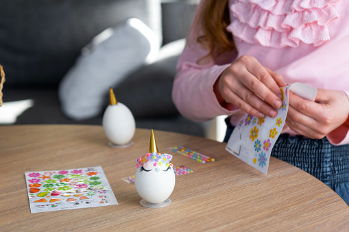 A cute girl with pink bunny ears makes an Easter craft - decorates an egg in the form of a unicorn with rhinestones, horn, flowers in the interior of a house with plants.