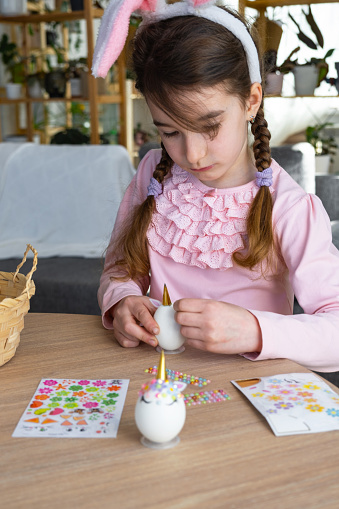 A cute girl with pink bunny ears makes an Easter craft - decorates an egg in the form of a unicorn with rhinestones, horn, flowers in the interior of a house with plants.