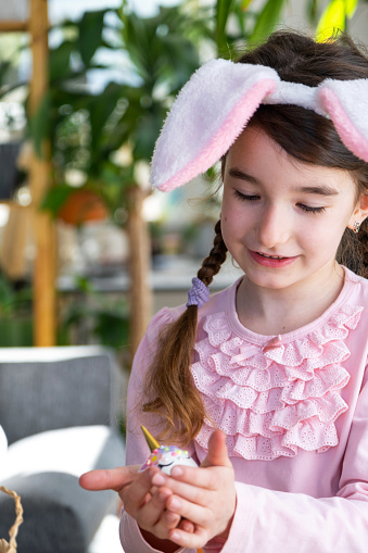 A cute girl with pink bunny ears makes an Easter craft - decorates an egg in the form of a unicorn with rhinestones, horn, flowers in the interior of a house with plants.