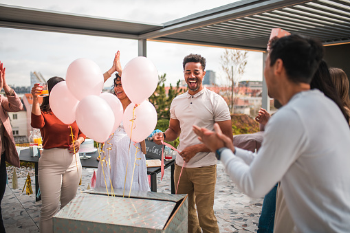 An excited, diverse group gathers outdoors for a gender reveal party, where pink balloons indicate a baby girl. People in casual attire are chatting and laughing, some capturing the moment on a smartphone amidst a decorated patio setting.