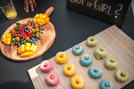 A variety of colorful donuts and fresh fruit platter on a table at a gender reveal party, with a 'Boy or Girl?' sign indicating a baby girl celebration. Indoors setting with a focus on celebration snacks.