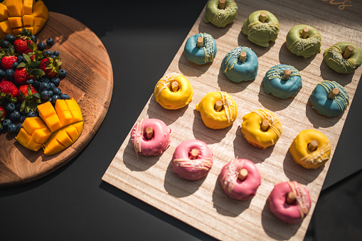 A selection of gourmet doughnuts in pink icing for a gender reveal party, denoting a baby girl. The treats are displayed elegantly alongside fresh berries and sliced mango, suggesting a cheerful celebration indoors.