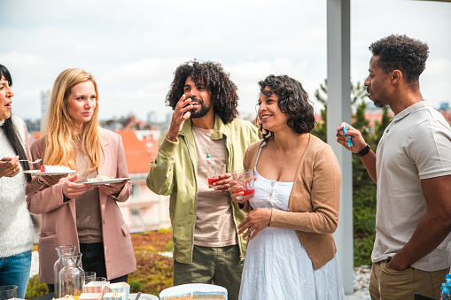 A mixed-race group enjoys a rooftop gender reveal party, with a pregnant woman in a white dress holding a drink. The casual gathering features delighted expressions, party food, and outdoor seating.