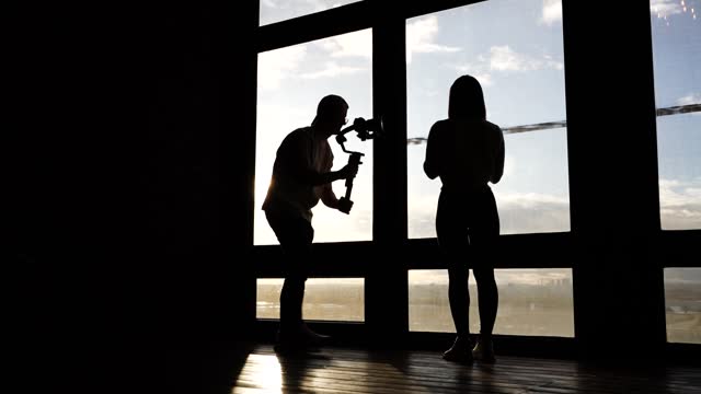 Silhouette of female model and male videographer near bright studio window