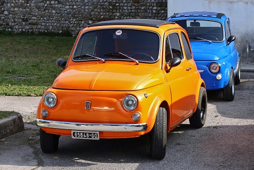 Percoto, Italy. March 17, 2024. Bright orange vintage Fiat 500 with another blue one behind, before a classic car parade.