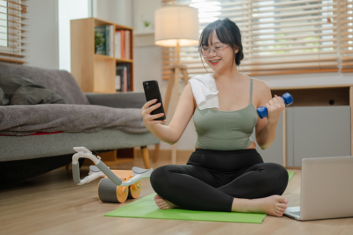 An attractive young Asian woman in sportswear is exercising at home, lifting a dumbbell and using her smartphone on a yoga mat.