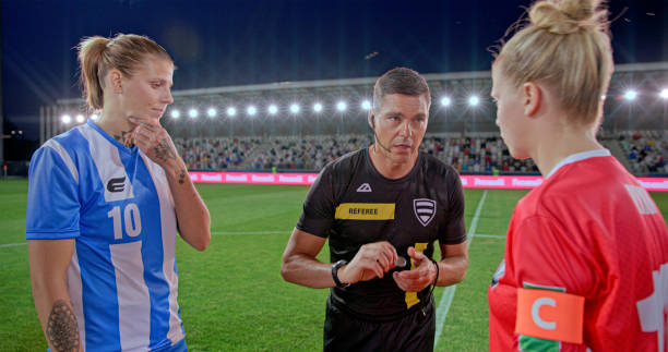 Referee tossing coin while standing with team captions during toss on football pitch.