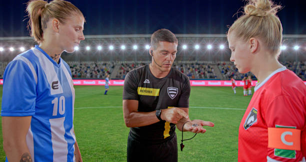 Referee tossing coin while standing with team captions during toss on football pitch.