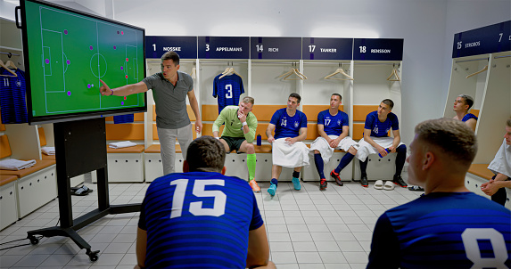 Football coach explaining game strategy on device screen to football players sitting in dressing room.