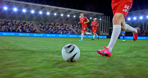 Low section of football player in red jersey gribbling ball during match at night.