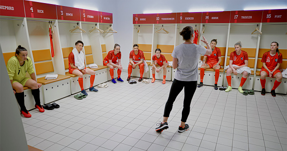 Female football coach gesturing while explaining game strategy to football players sitting in dressing room.