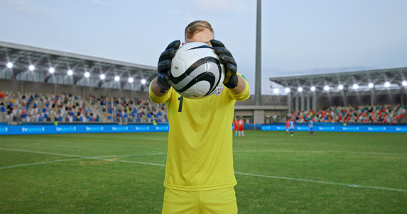 Male goalkeeper in yellow jersey standing while holding football on pitch during match.