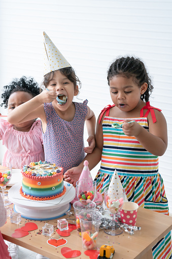 Group of multiracial children friends enjoy eating rainbow cake decorated with sugar candies and jelly on birthday party with spoon. Children have fun celebrating party at home with mess up mouth