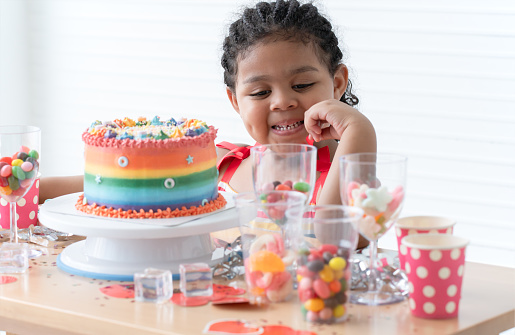 African little kid girl in cute dress, surprised on birthday party with beautiful rainbow cake decorated with sugar candies, colorful sprinkles. Colorful candies, jelly in glasses and hats on table