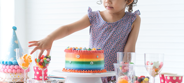 Selective focus on hand of little kid girl use finger to pick up candies from glass, happy child on birthday party with beautiful rainbow cake decorated with sugar candies, colorful jelly on table