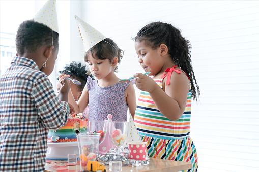 Group of multiracial children friends wear hat enjoy eating rainbow cake, sugar candies and jelly on birthday party with spoon. Children have fun celebrating party at home with mess up mouth
