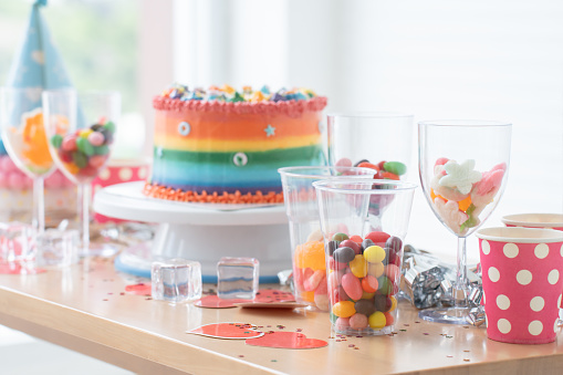 Focus on colorful candies in glass. Cake on birthday with rainbow butter cream on white background decorated with sugar candy. Cute cake placed on stand with jelly, hats on table. Festive celebration