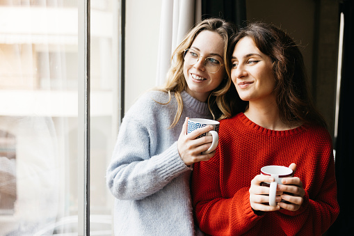 Two youthful women standing side by side, holding a cup of tea, near a window inside their home, sharing a moment of companionship in an intimate and cozy atmosphere.
