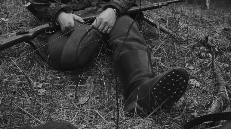 Reenactor Dressed As German Infantry Soldier Sit On Ground With Carabin Mauser 98k In Hand. Details Of Marching Boots Marschstiefel. Close-up View On Wermacht German Military Uniform During Ww2. Black And White Video