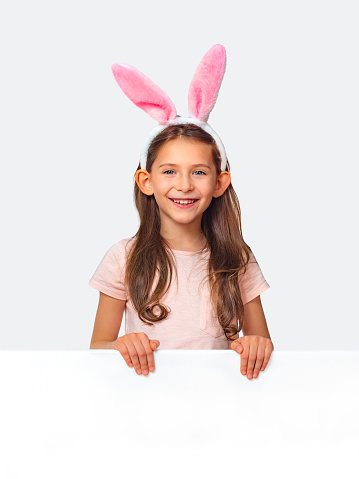 Cheerful girl in bunny ears peeks out from behind a sheet of paper isolated on a white background. A girl hunts for Easter eggs.