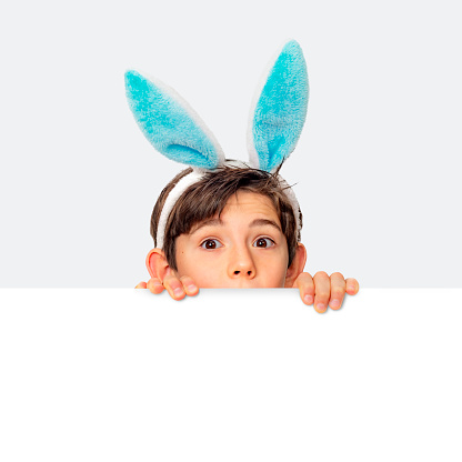 A boy hunts for Easter eggs. A boy wearing bunny ears peeks curiously from behind a white piece of paper. Isolated on a white background.