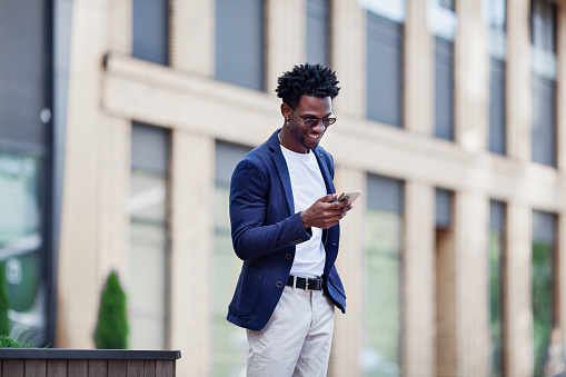 Stylish African American man in smart casual clothes, with mobile phone against backdrop of city. Fashionable guy with smartphone outdoors.