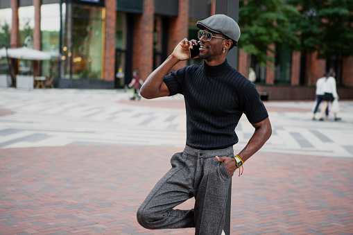 Handsome stylish black man in a cap and black t-shirt emotionally engages in a phone conversation and smiles outdoors in the city against a building backdrop.